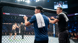 Two athletic cornermen watch the octagon cage under bright arena lights, towels draped on shoulders