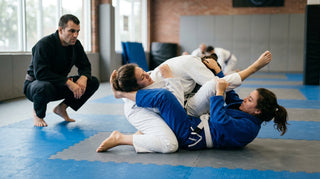 Two gi-clad grapplers mid-roll on mats, coach observing closely from the edge