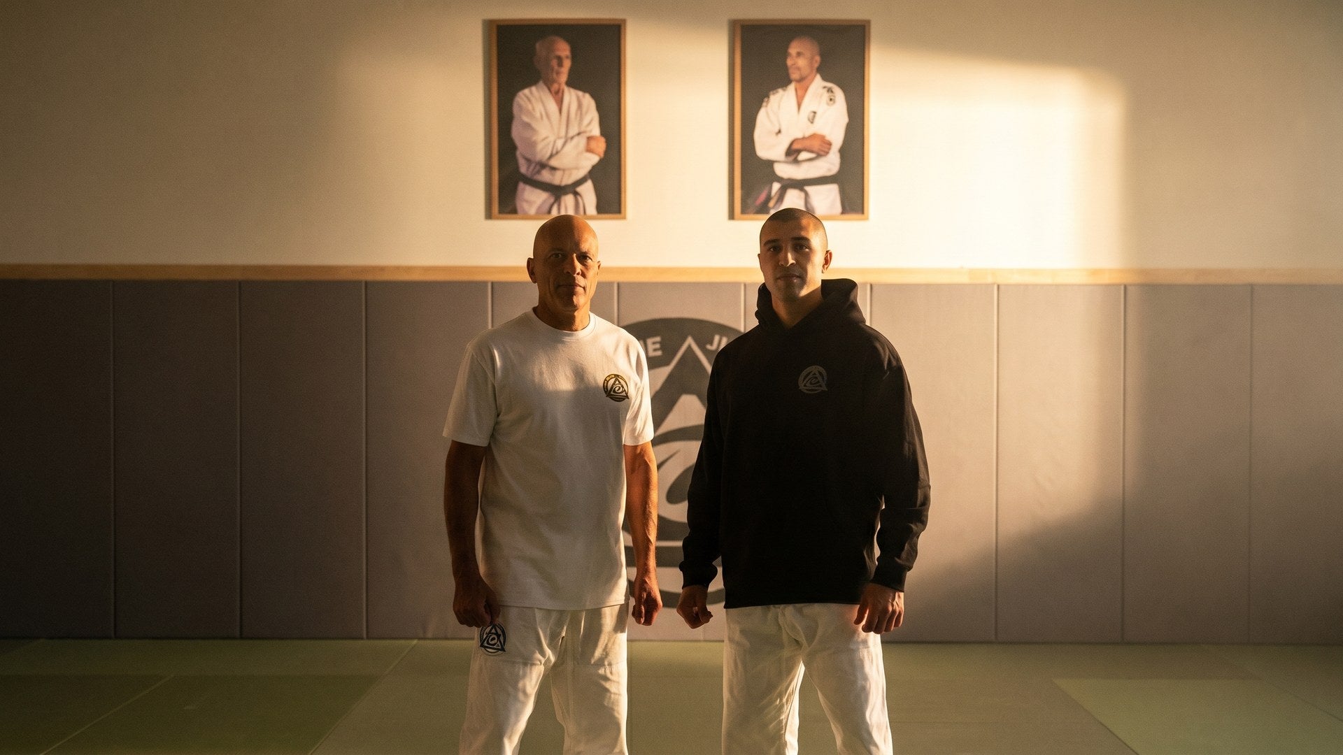 Royce Gracie and Khonry Gracie in family dojo with Helio Gracie and Carlos Gracie lineage portraits behind them, wearing Network Early Access Academy 360 Tee and Signature Hoodie, dramatic heritage lighting